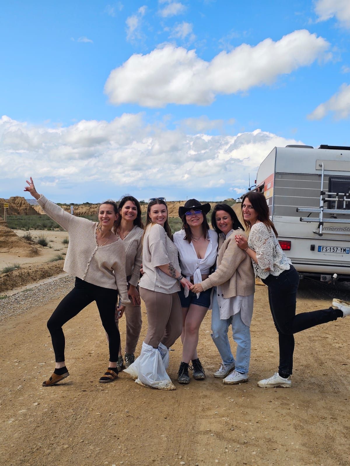 A group of friends poses playfully in front of a scenic desert backdrop with a parked RV trailer in the distance.