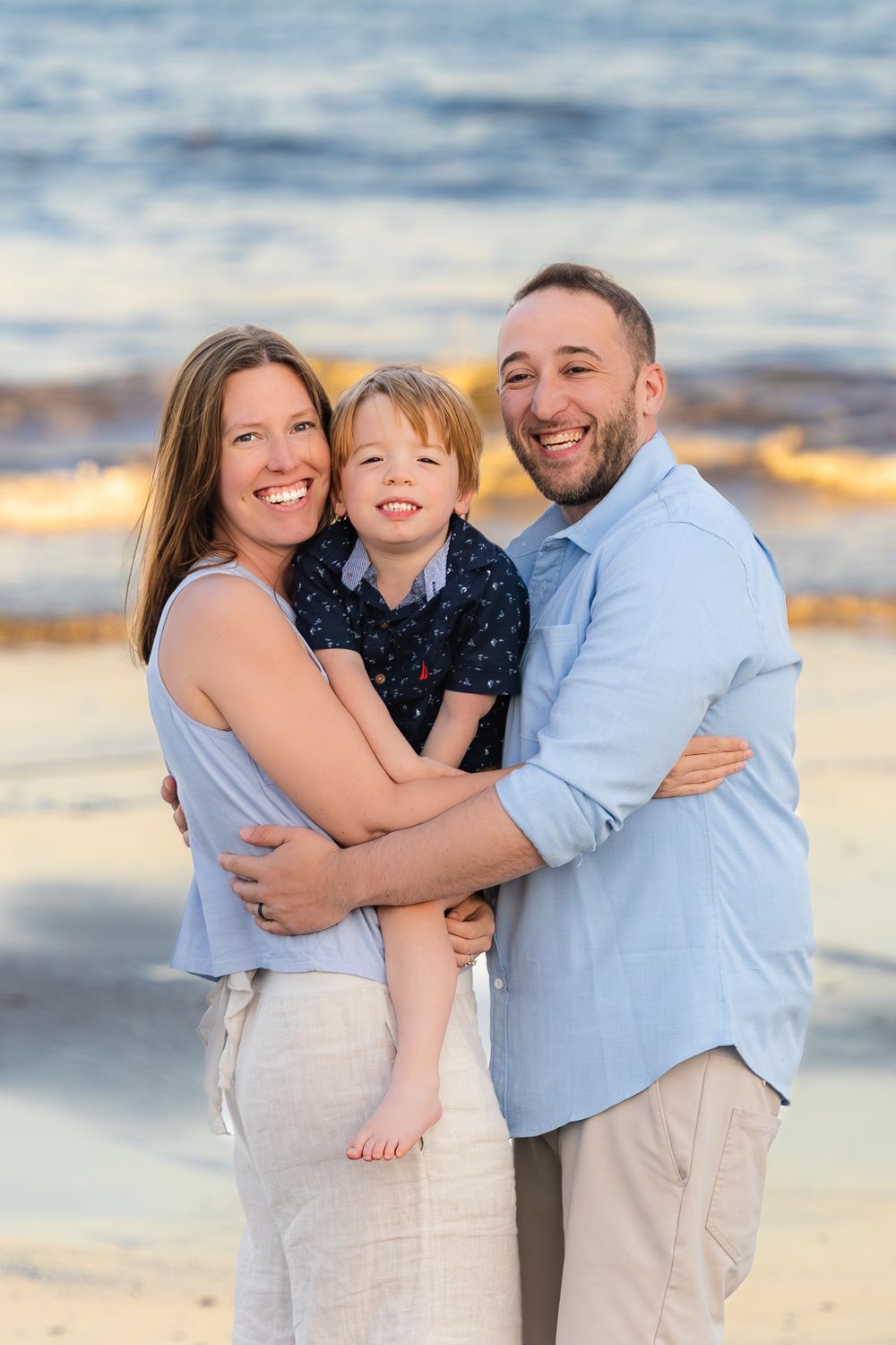 Family embraces together on a beach at sunset wearing coordinated light blue and white outfits.