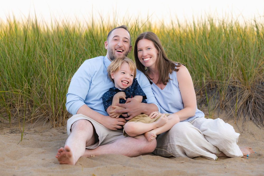 A happy group relaxes and poses together on a sandy beach during sunset with tall grass in the background.