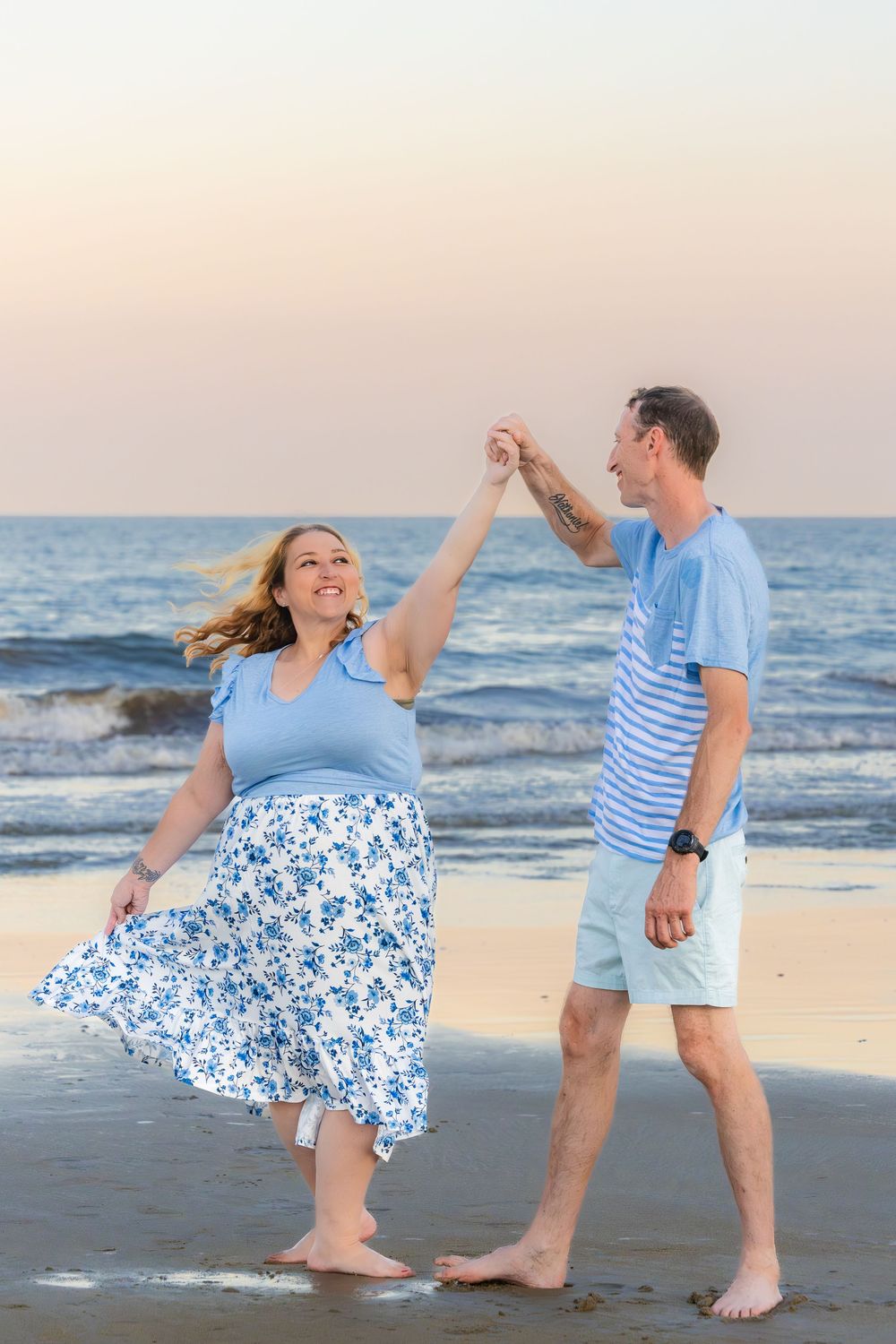 A couple dances and twirls together on the beach at sunset wearing coordinating blue and white outfits.