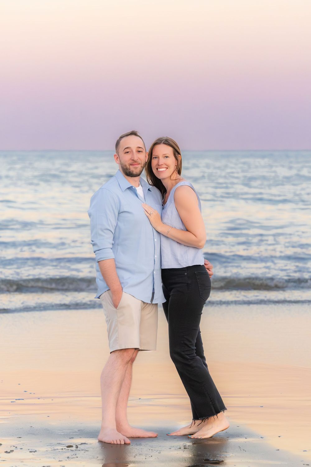 A couple shares a romantic moment together barefoot on the sandy beach during a pink and purple sunset.