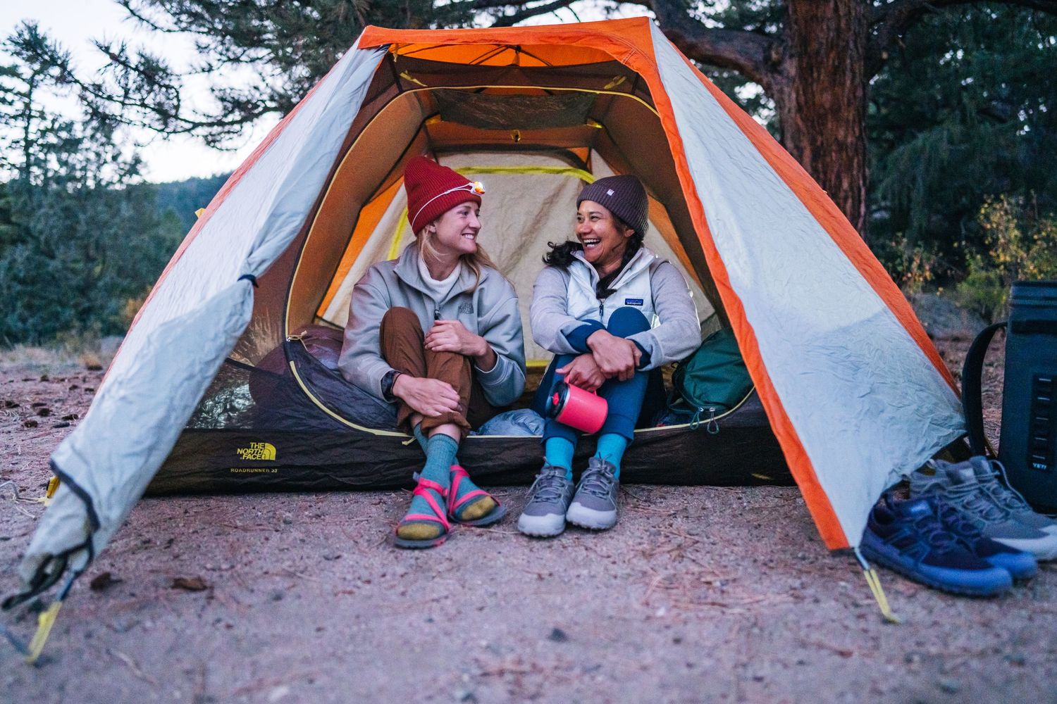 Two campers sit and chat inside an orange tent while enjoying coffee during a peaceful outdoor adventure.