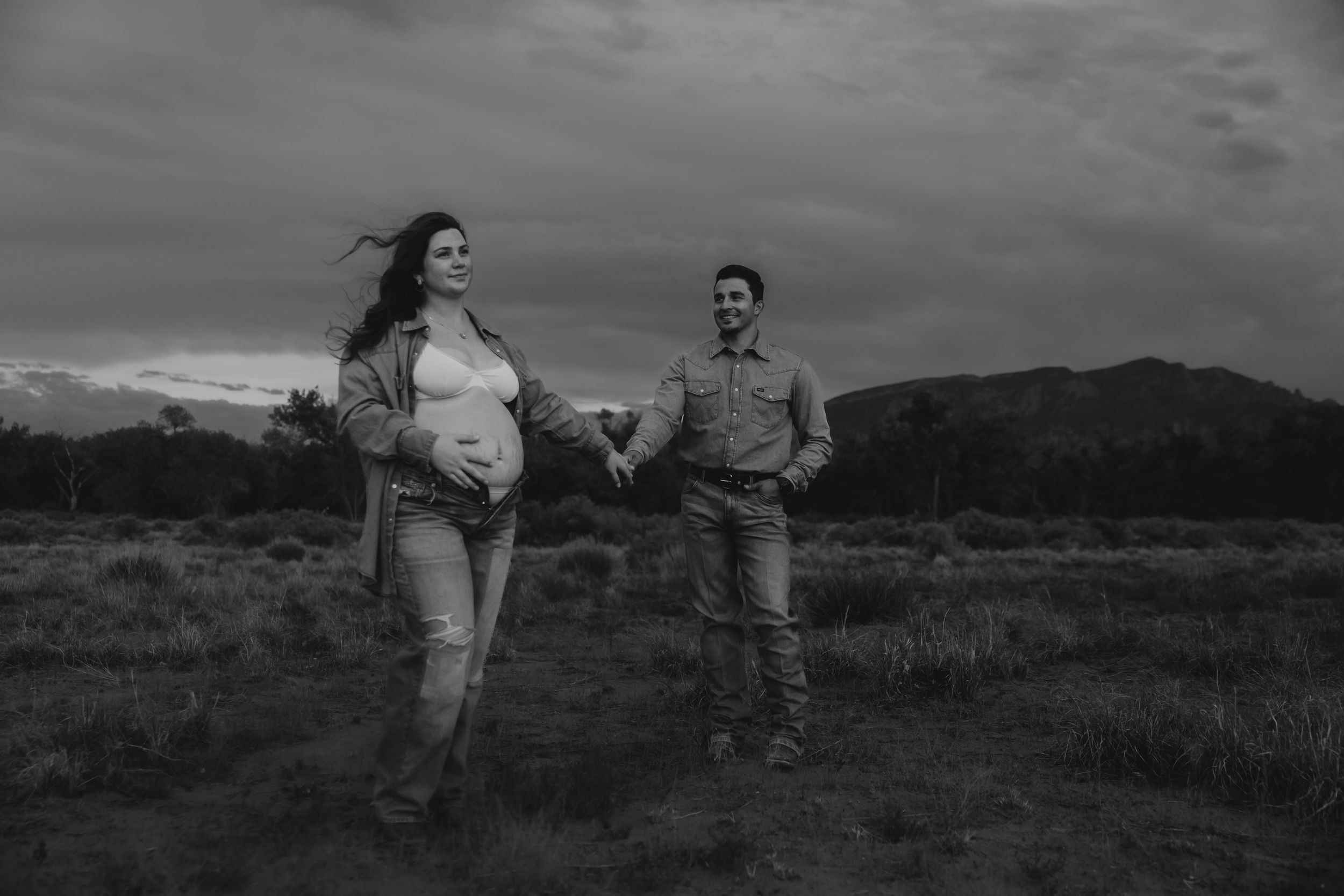 Black and white maternity photo of couple holding hands in open field with mountains.