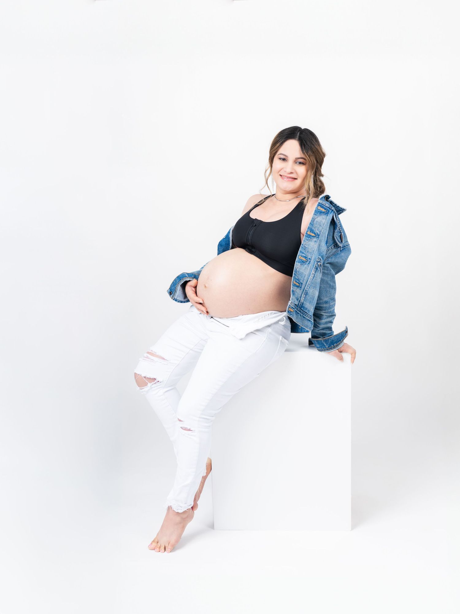 Maternity photo shoot featuring casual denim jacket and white pants against bright white background.