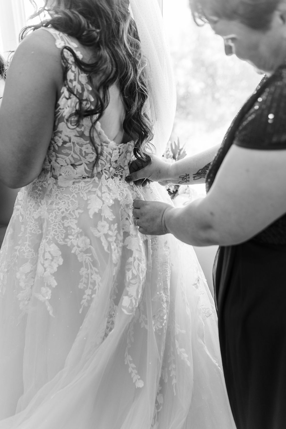 A detailed black and white photo showing someone helping adjust the back of an ornate lace wedding dress.