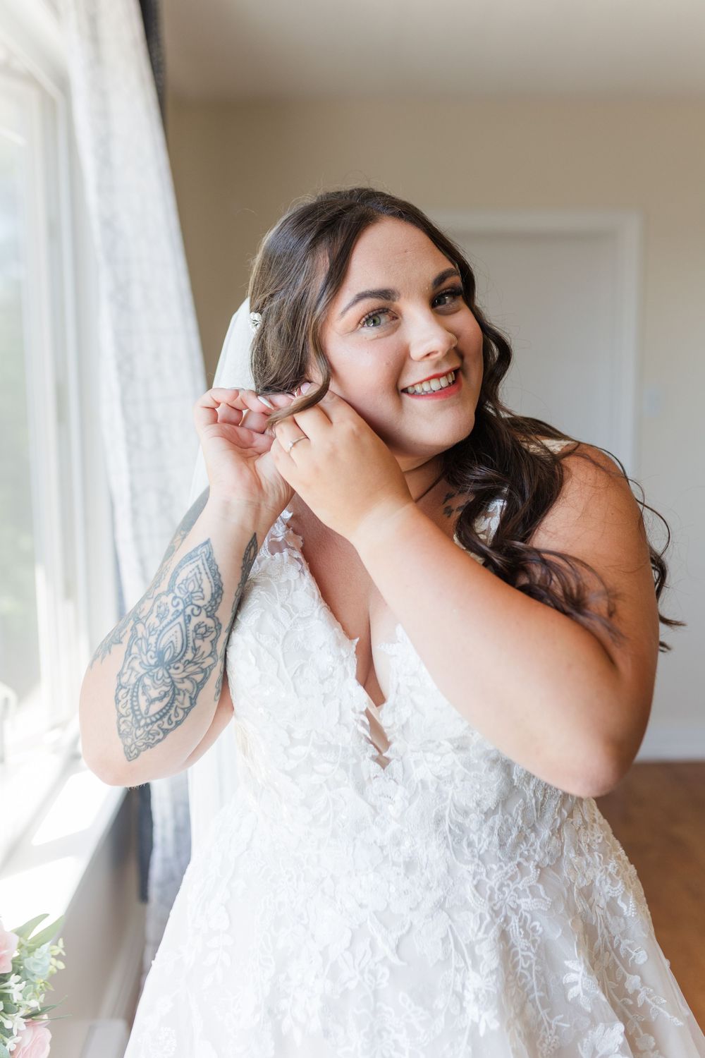 Someone in an elegant white wedding dress with lace details and dark wavy hair smiles joyfully while getting ready.