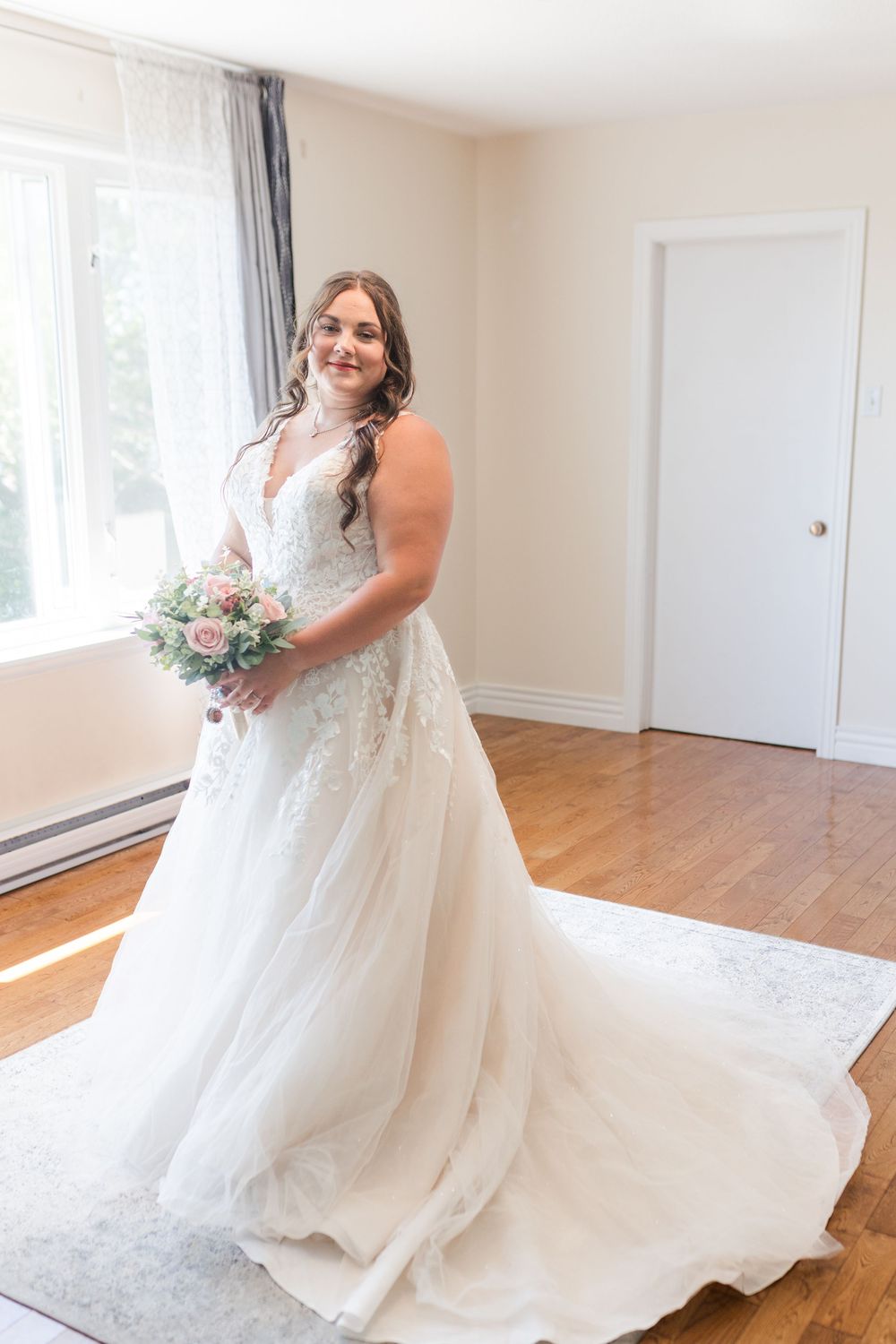A bride in a beautiful white lace gown with a long train holds a pink and white bouquet in an elegant sunlit room.