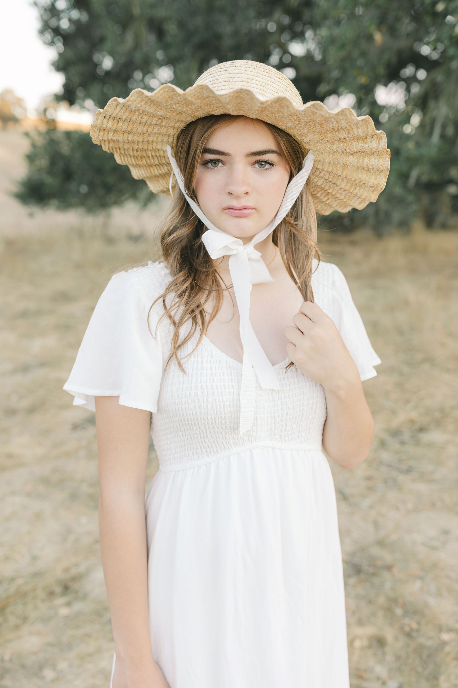 Woman in white dress stands in golden field at sunset posing gracefully with flowing fabric and gentle breeze.Film by Chelsee Rawe Photograp