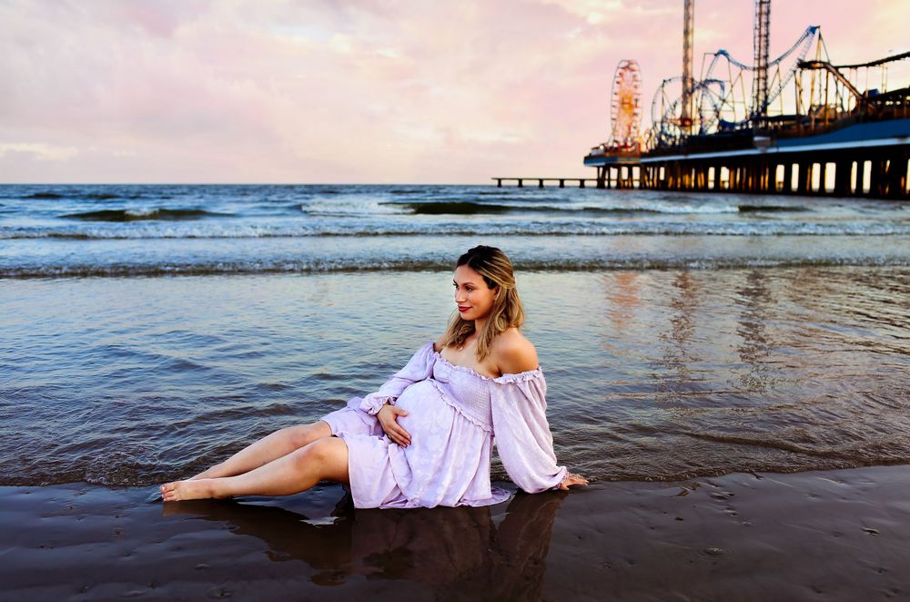 Pregnant mama in purple dress laying near the beach water while posing for a maternity session while holding her belly