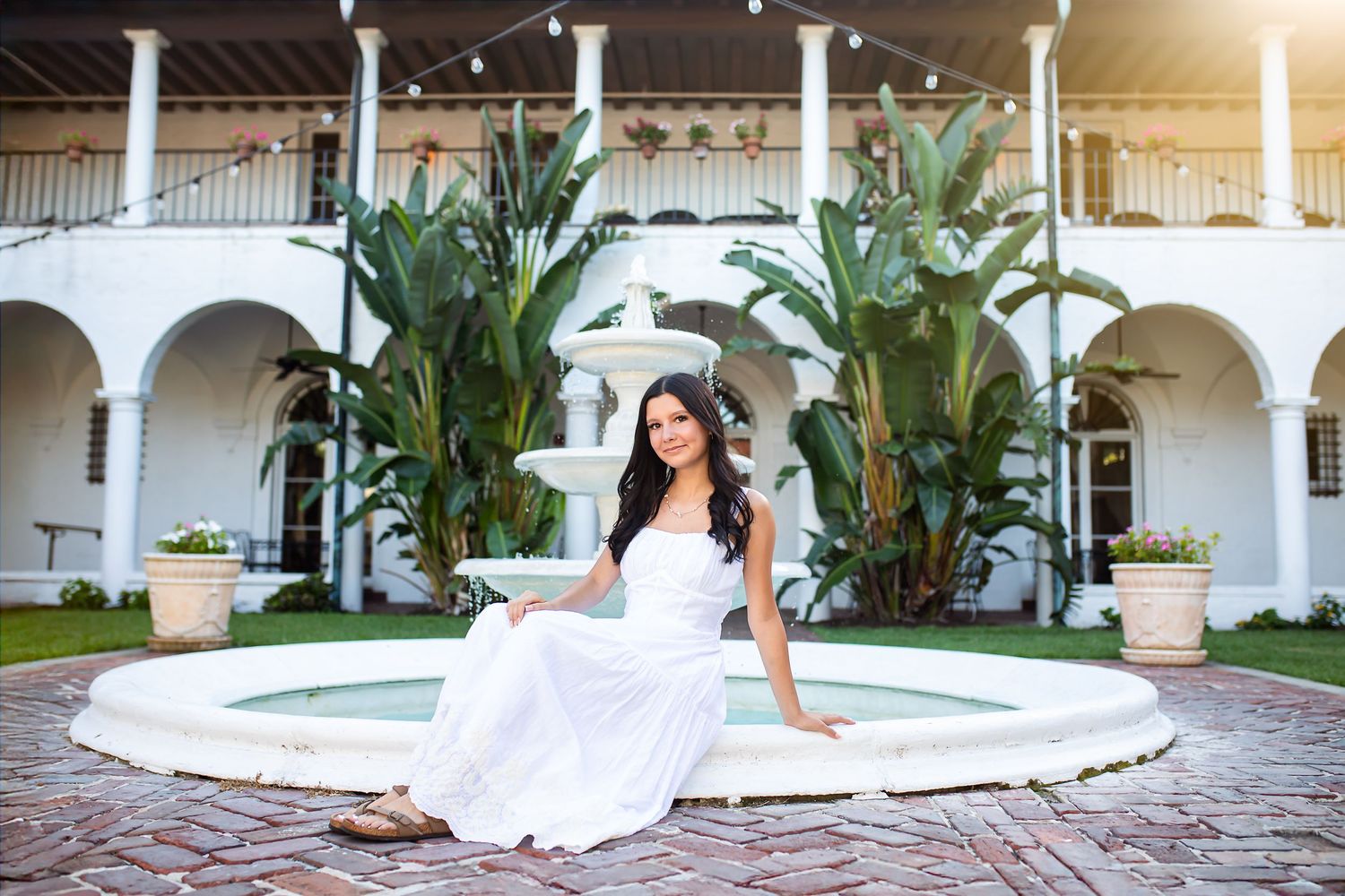 A wedding dress glides gracefully across a rustic brick courtyard with palm trees and white columns in the background.