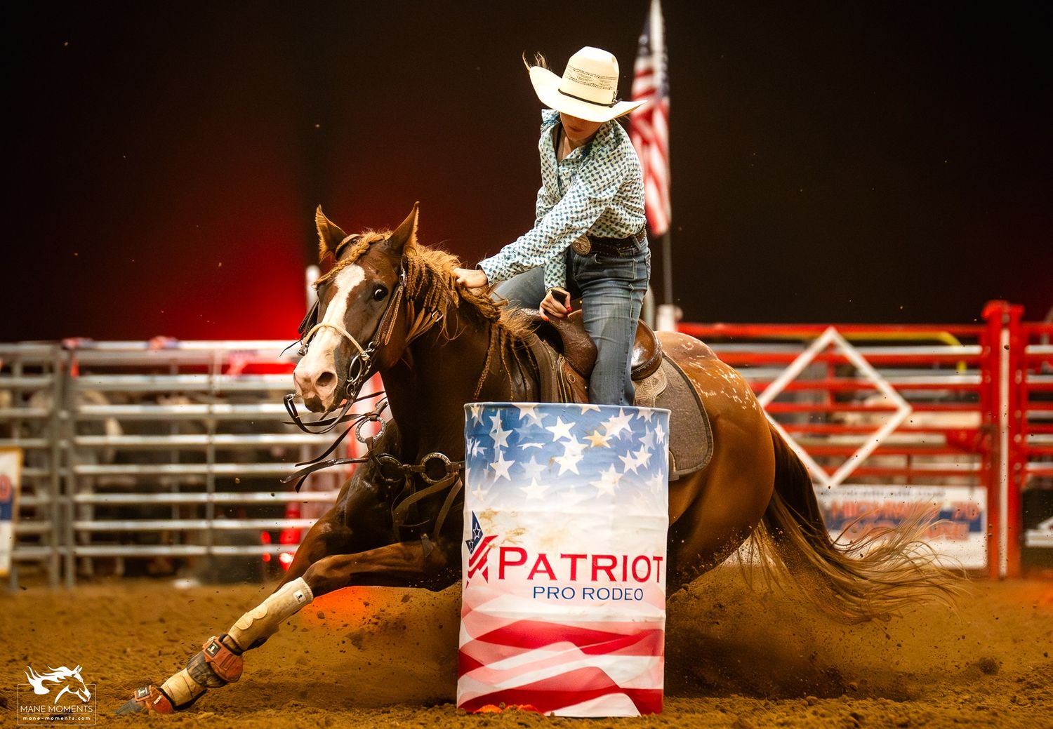 A rodeo rider on horseback rounds a patriotic red white and blue barrel in an illuminated arena during competition.