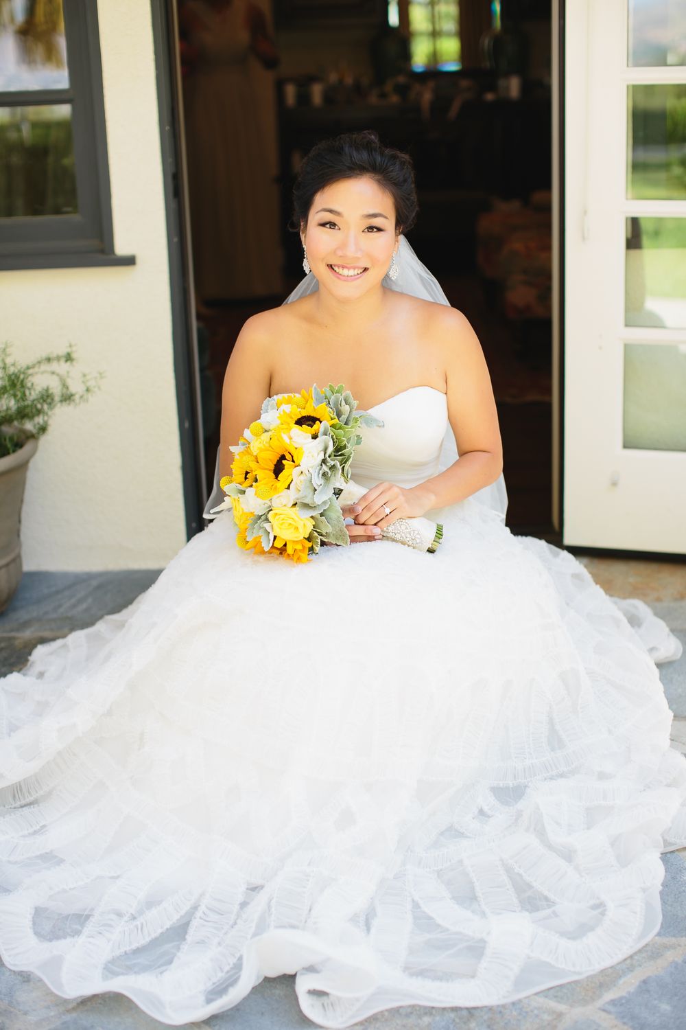 Bride in white wedding gown poses with bright yellow sunflower bouquet against light exterior background.