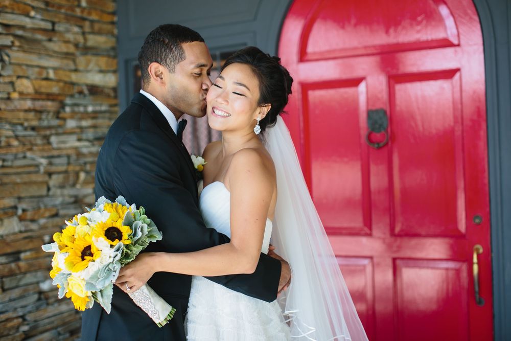 Wedding couple embraces near a striking red door and stone wall.