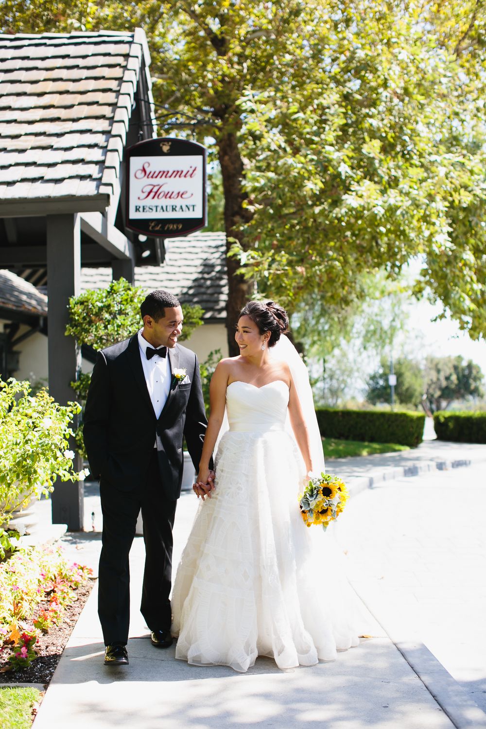 Wedding couple walks together along garden pathway under shady trees.