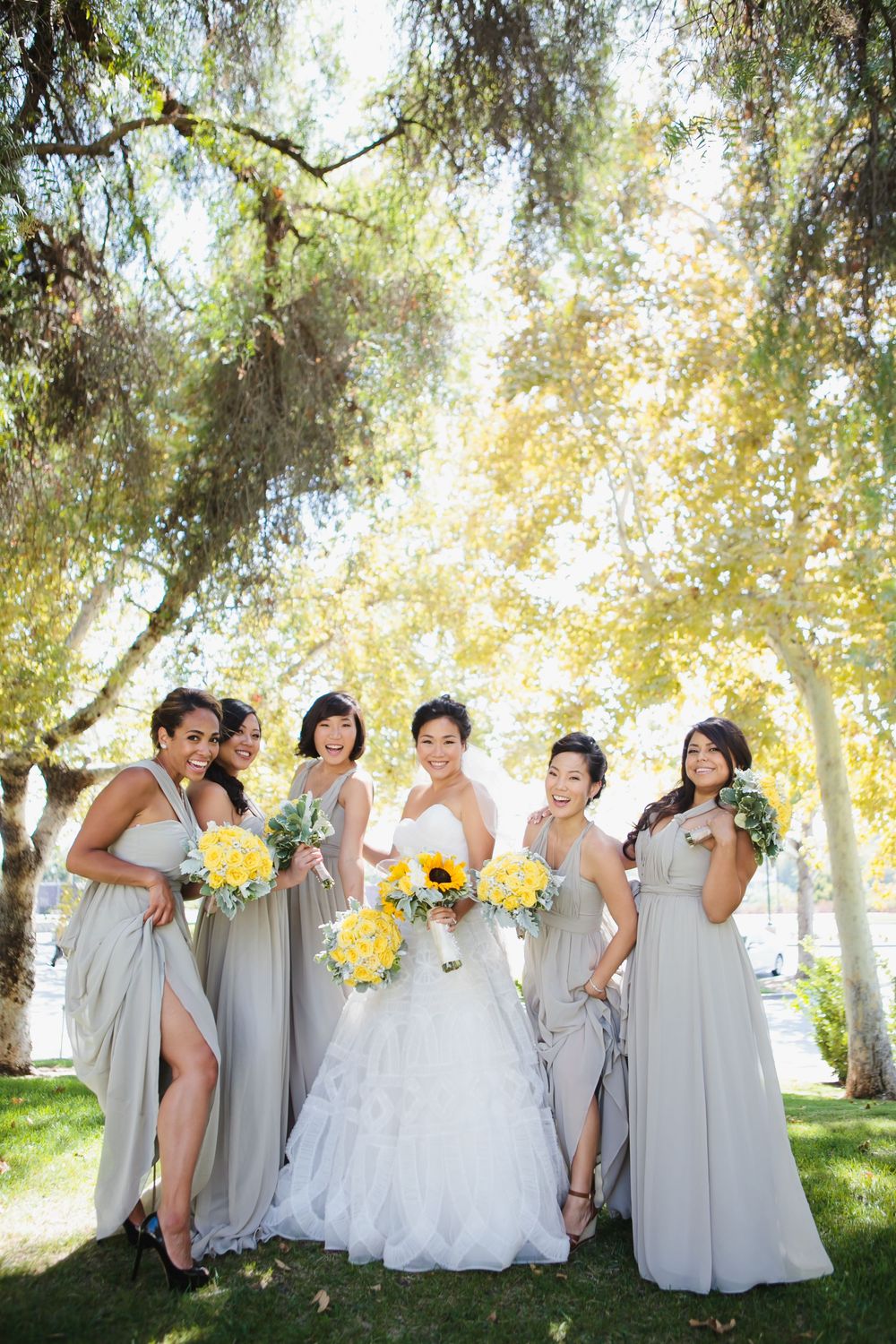 Bridal party in light grey dresses poses playfully with bouquets under willow trees.