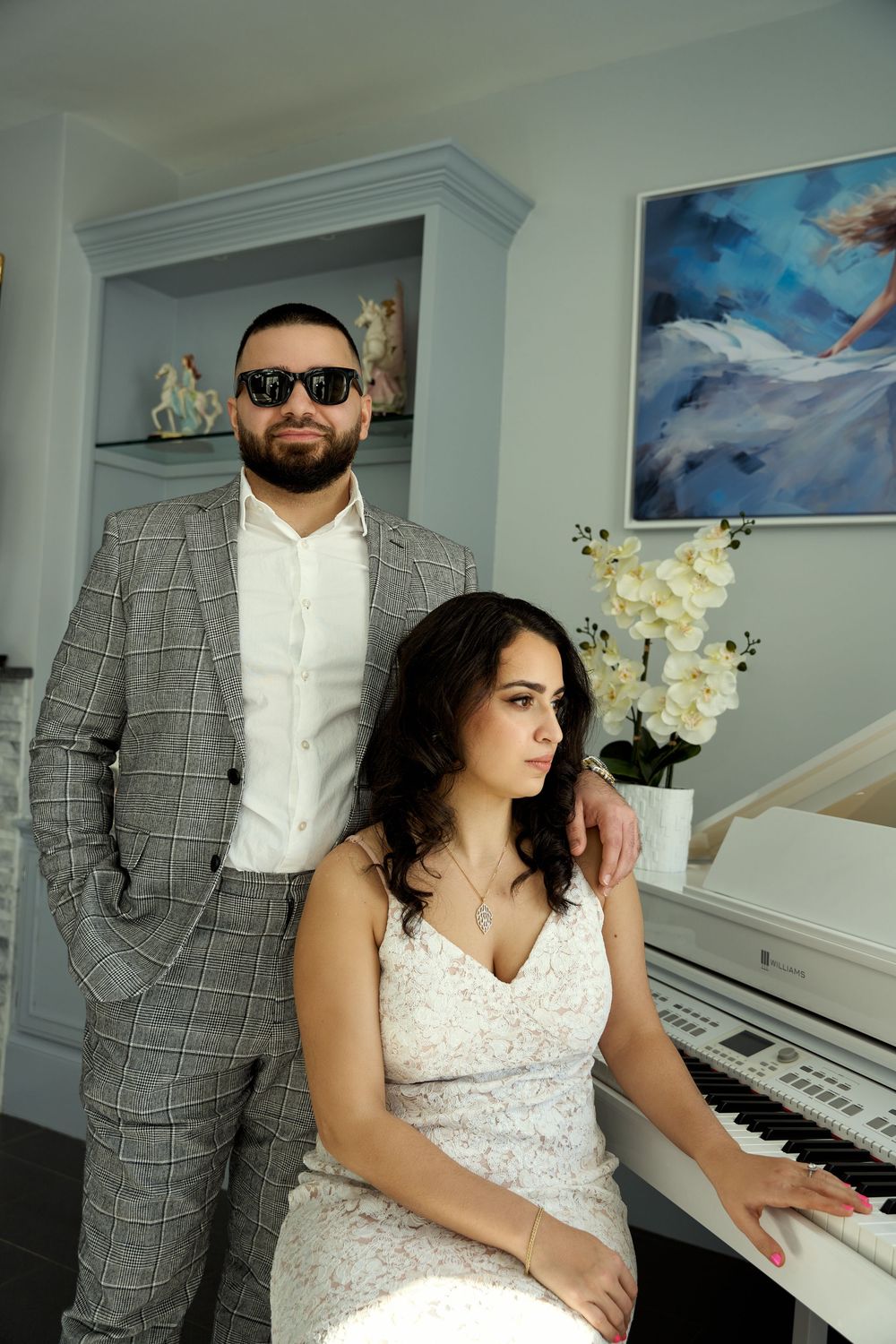 A couple in formal attire pose near a white piano with a blue abstract painting and orchids in the background. Engagement photoshoot