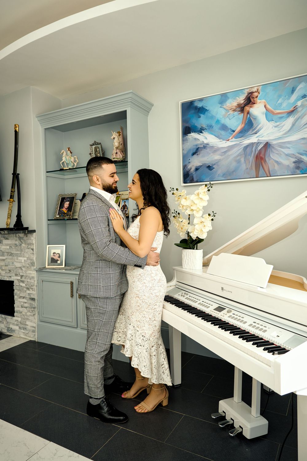 A couple dances together in a room with a white piano and light blue walls decorated with artwork. Engagement photoshoot