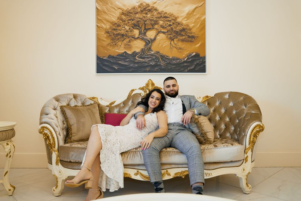 Two people sit together on an ornate gold and cream colored tufted sofa in a bright interior setting. Engagement photoshoot