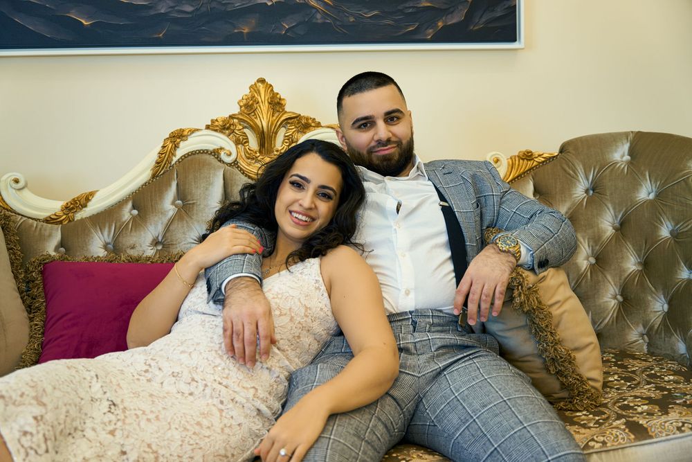 A couple relaxes together on an ornate gold and gray tufted sofa with decorative pillows. Engagement photoshoot