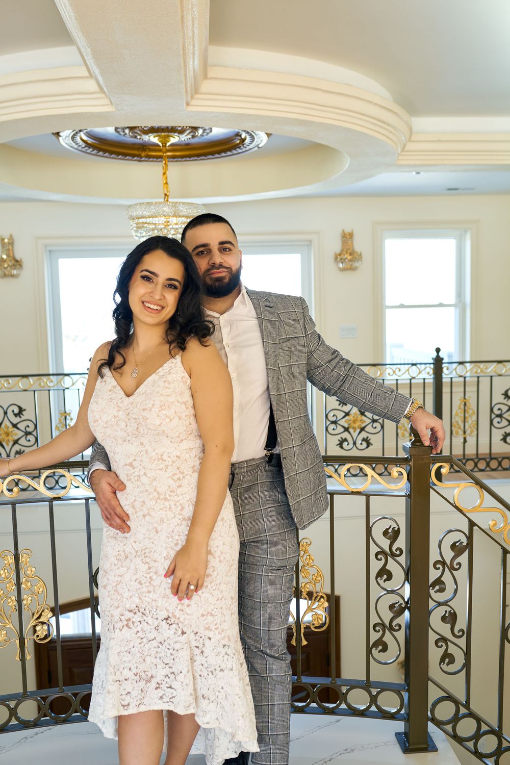 A couple poses elegantly on an ornate balcony with decorative railings and a grand ceiling medallion in an upscale interior. Engagement phot