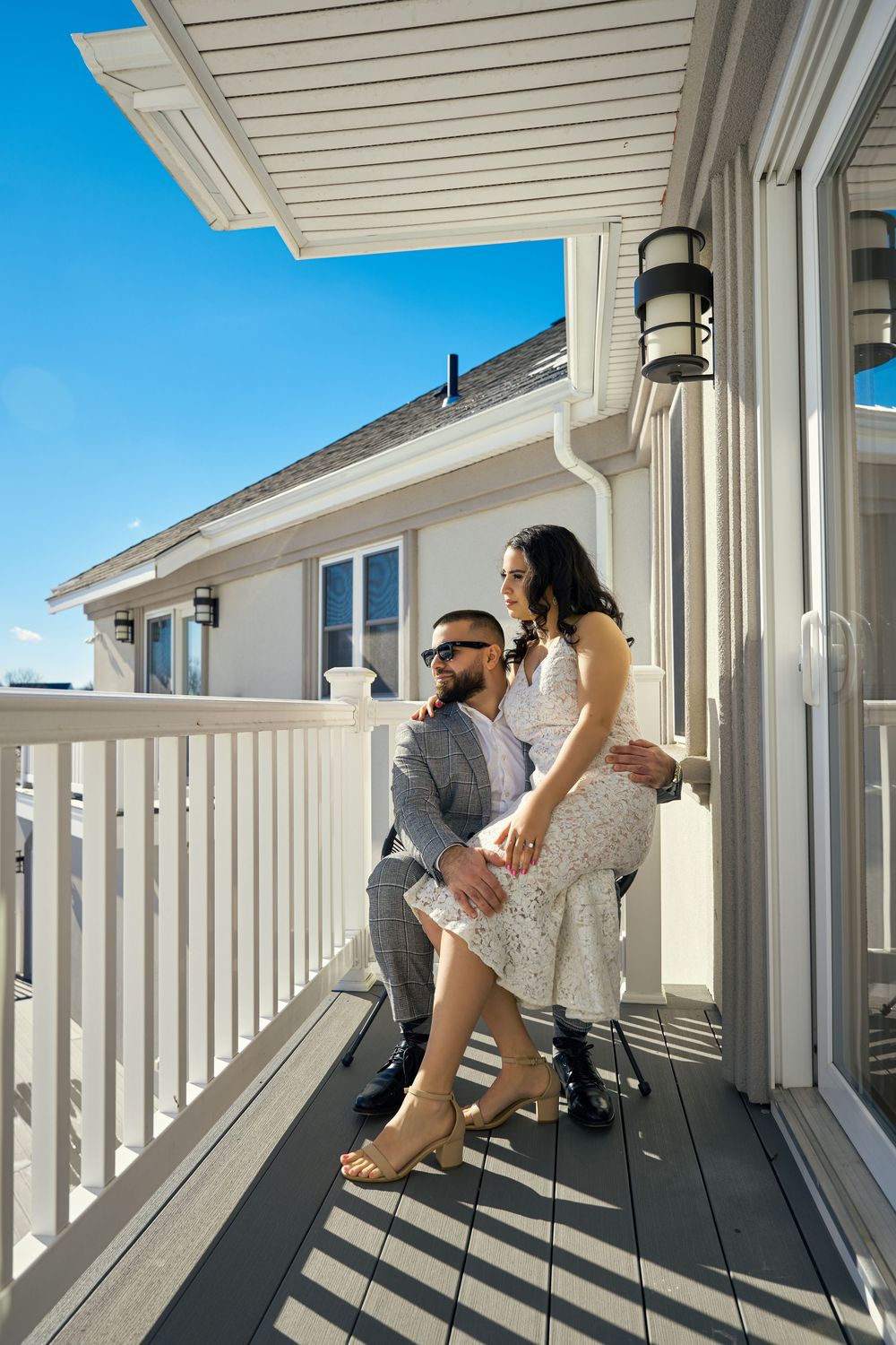 A couple shares an intimate moment on a white deck balcony of a coastal home on a bright sunny day. Engagement photoshoot