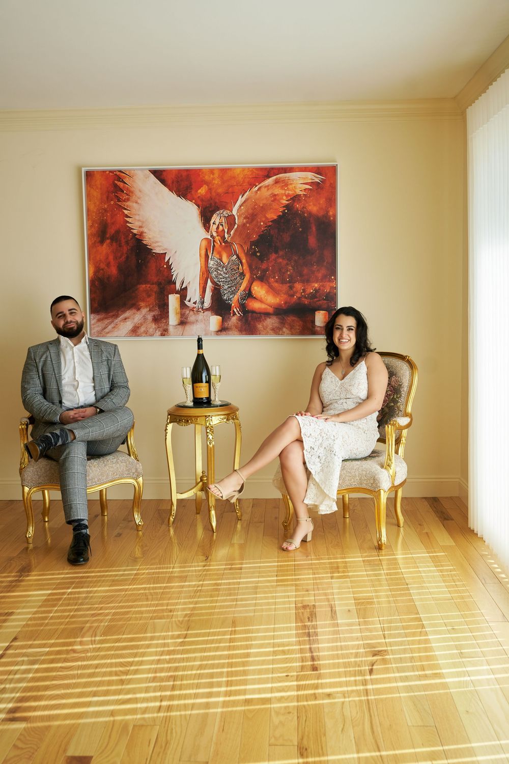 Two people seated in antique chairs with champagne and striking red angel artwork on the wall behind them. Engagement photoshoot
