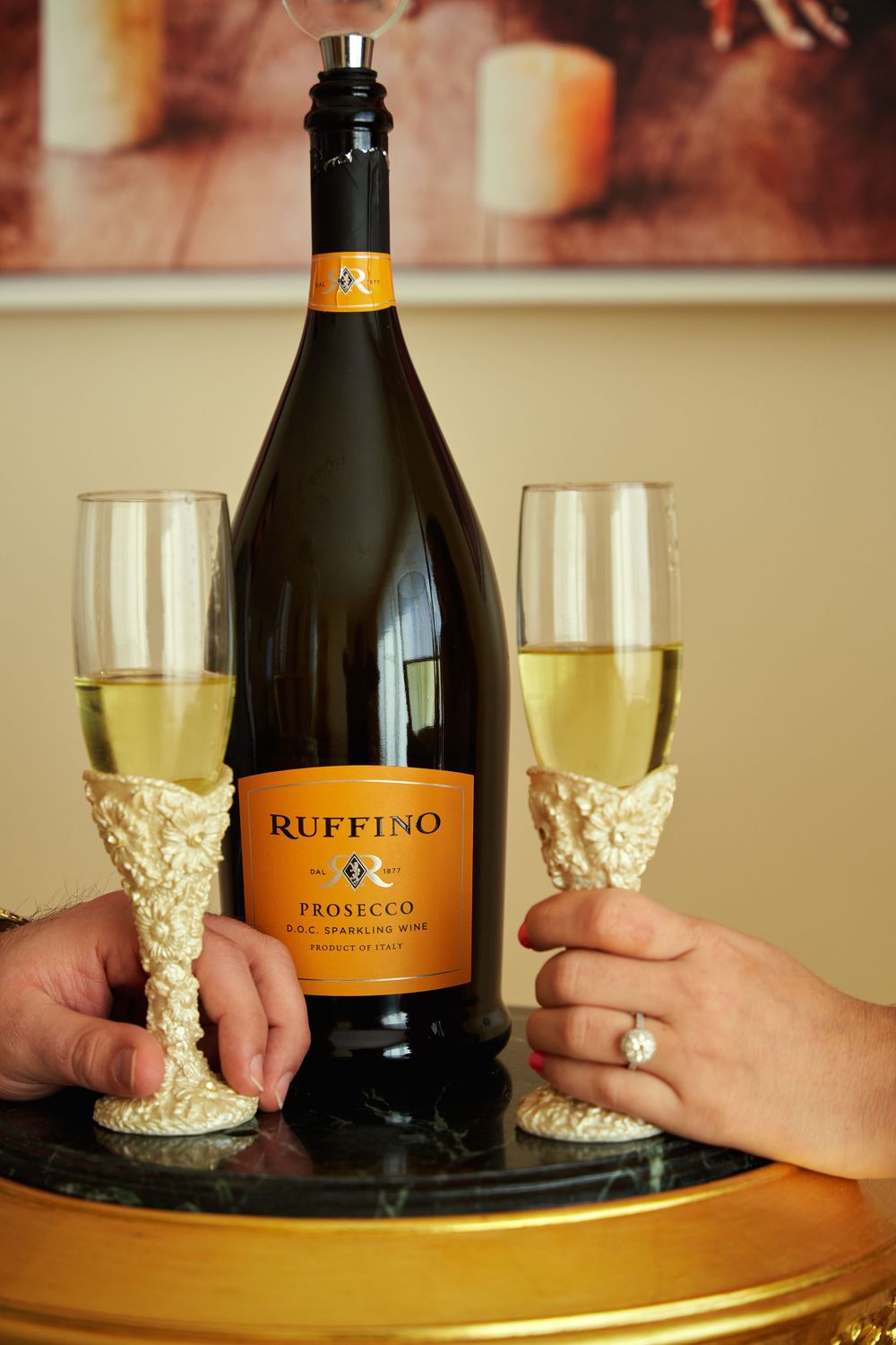 Two hands holding decorative champagne glasses with a bottle of Ruffino Prosecco wine on a round wooden table. Engagement photoshoot