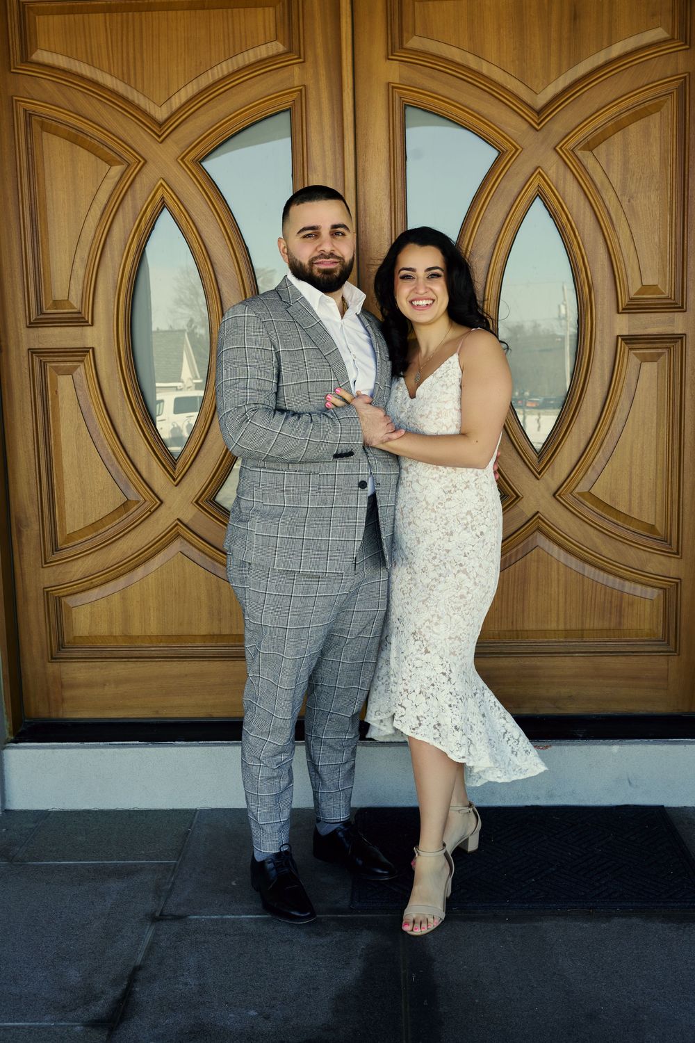 A couple stands together in front of wooden double doors with circular window details in formal attire. Engagement photoshoot