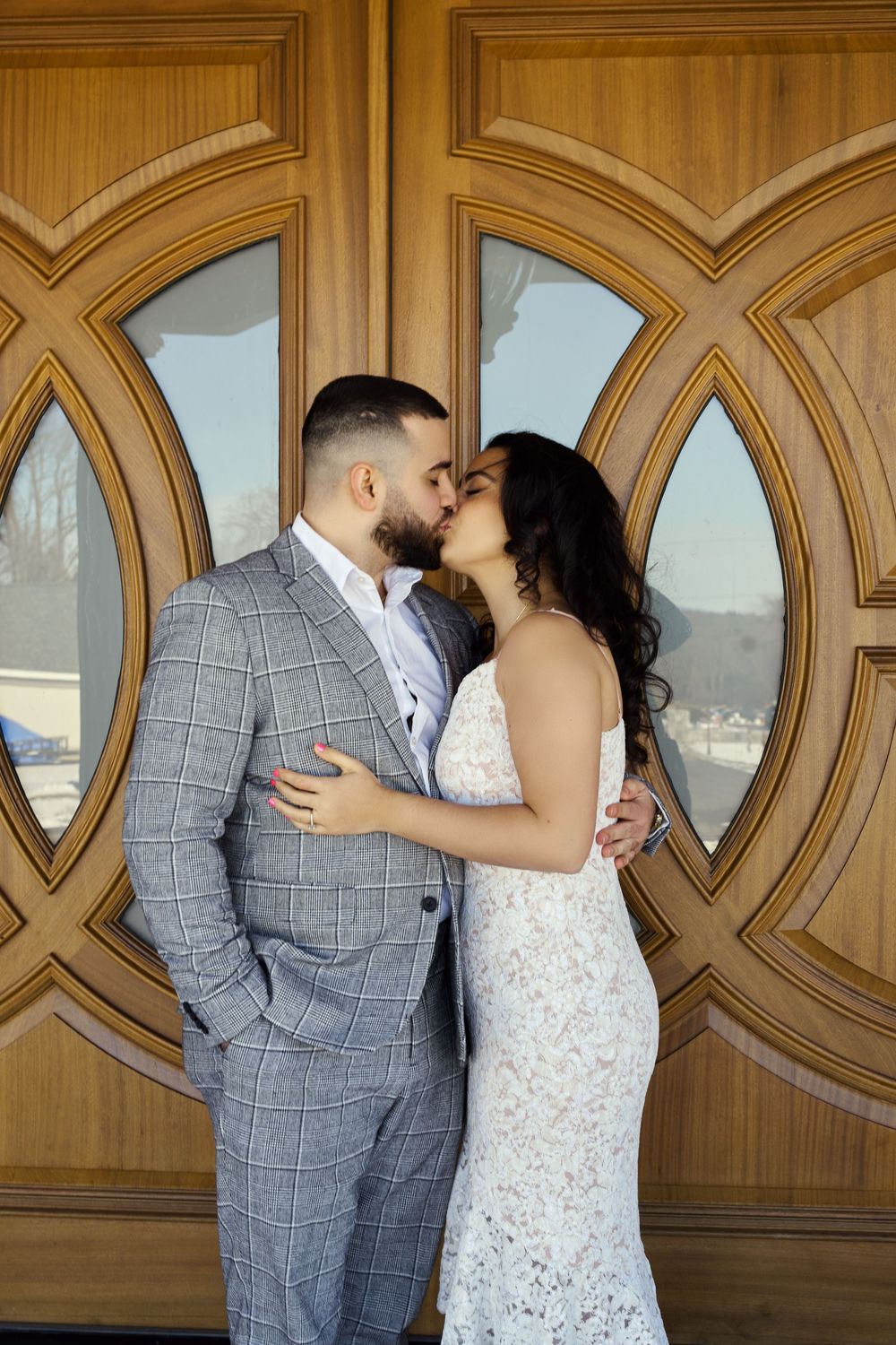 A romantic couple shares a tender kiss in front of ornate wooden doors with oval-shaped windows overlooking a scenic view. Engagement photos