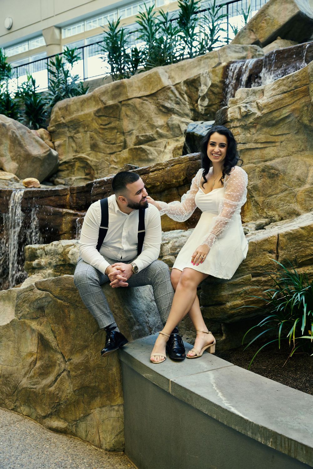 A couple in formal attire poses together on decorative stone rocks with a waterfall feature in an indoor setting. Engagement photoshoot