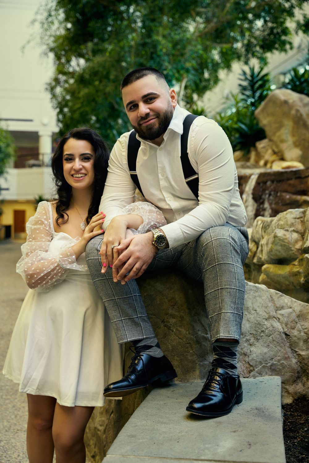 A couple poses together on rock formations outdoors, with her in a white dress and him in dress pants and vest. Engagement photoshoot