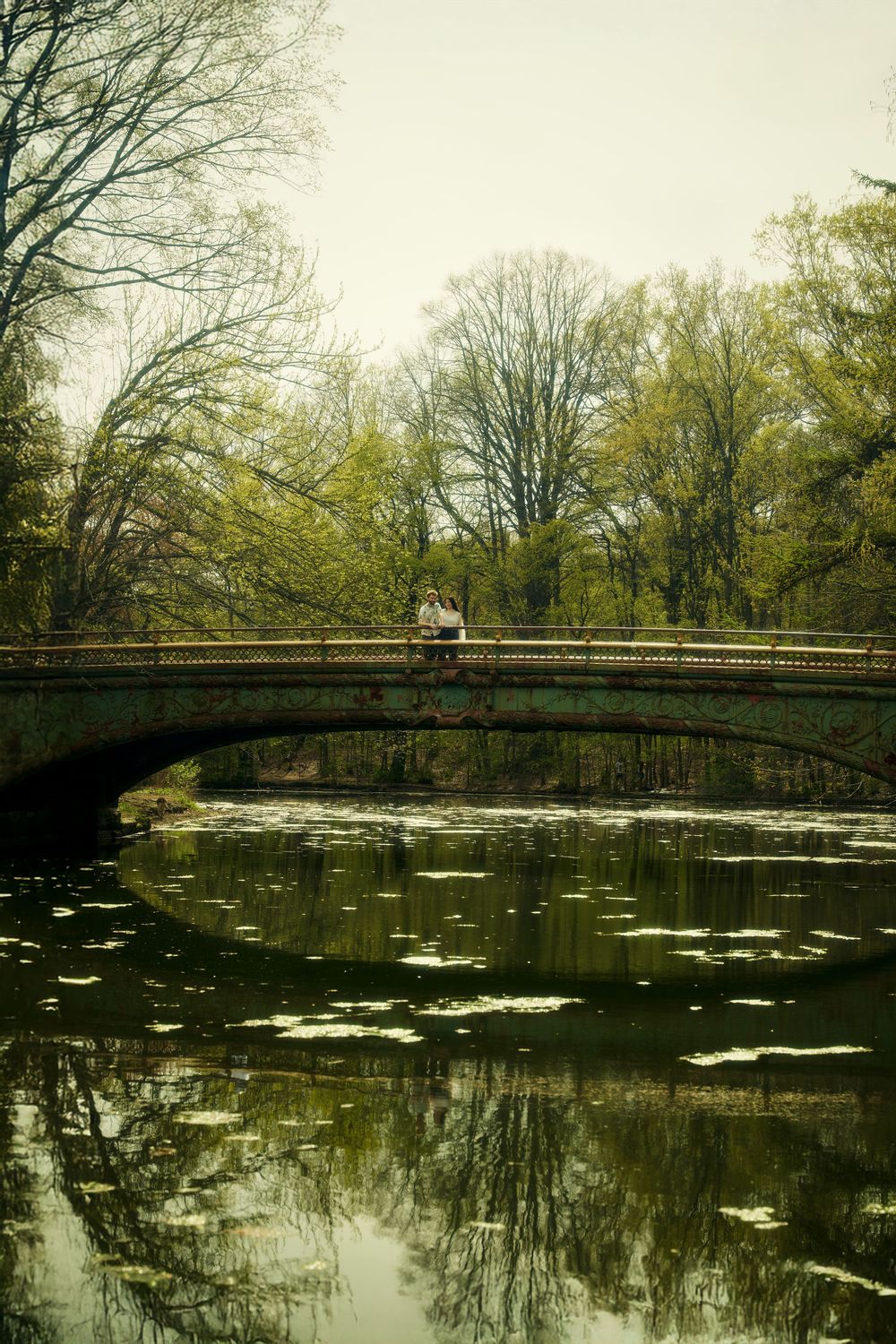 Two people stand on a wooden footbridge over a tranquil pond with lily pads and tree reflections in the water. engagement photoshoot