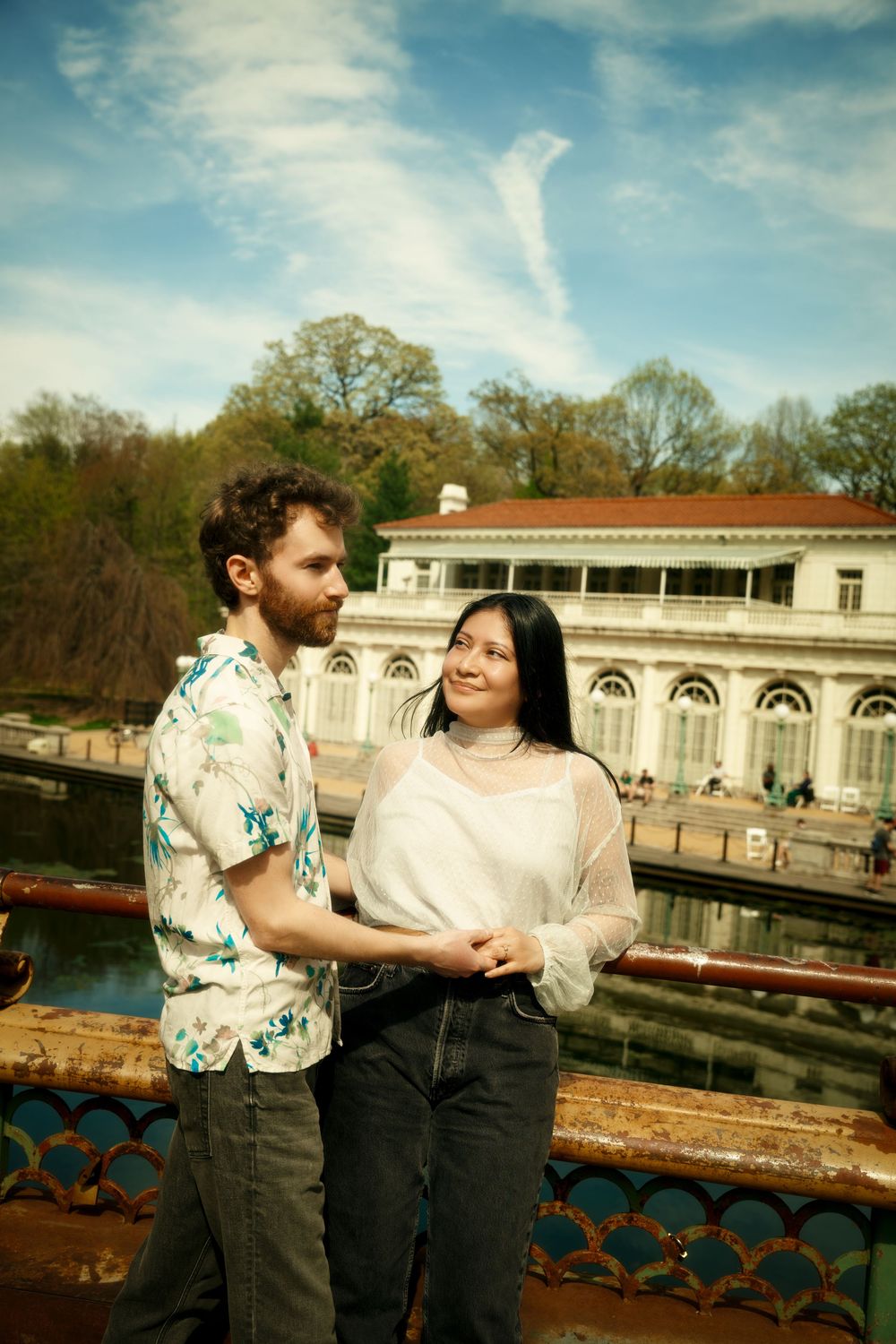 A couple shares a romantic moment overlooking a historic mansion with a reflecting pool on a sunny day. at prospect park boat house