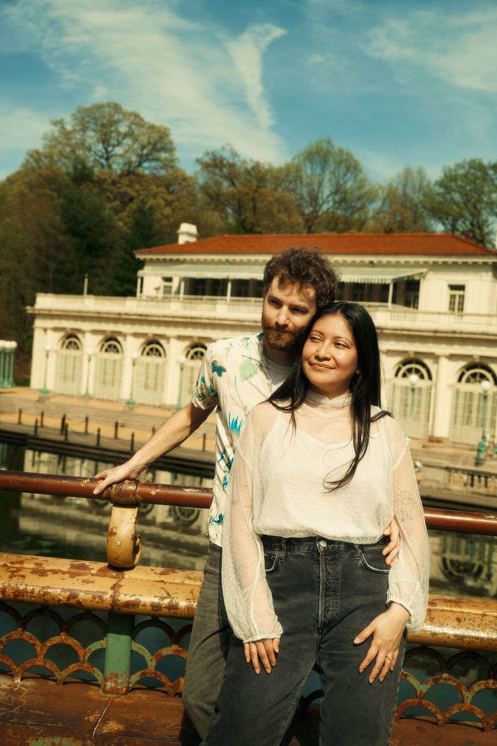 A couple stands together on a terrace with ornate architectural details and trees visible in the background. at prospect park boat house