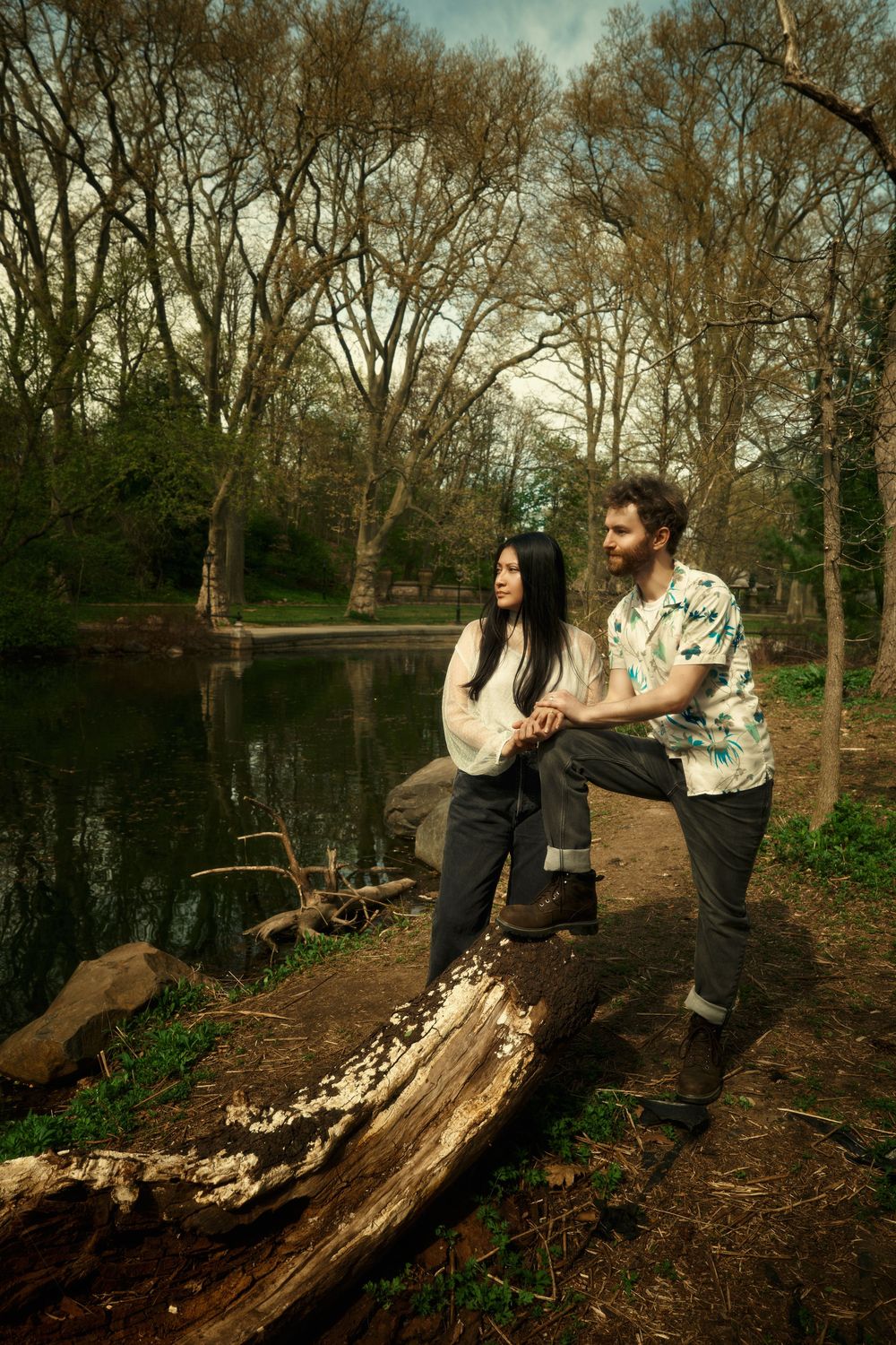 A couple poses together on a fallen log by a serene lake in a wooded park during autumn at prospect park boat house