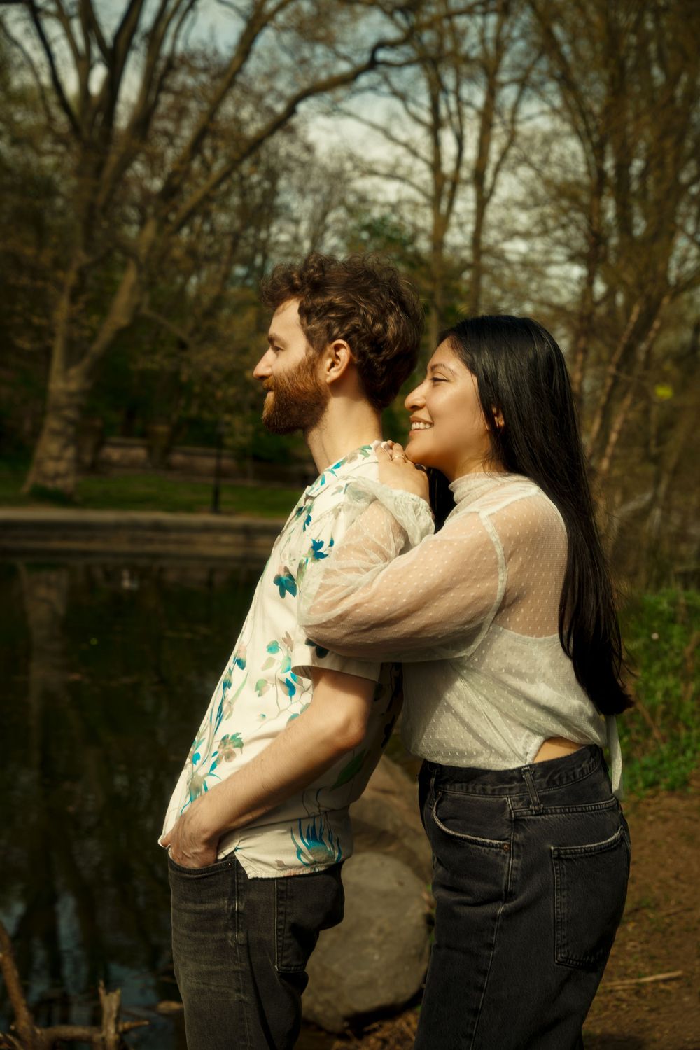Two people share a joyful moment together while standing by a pond in a wooded area on an autumn day. engagement photoshoot