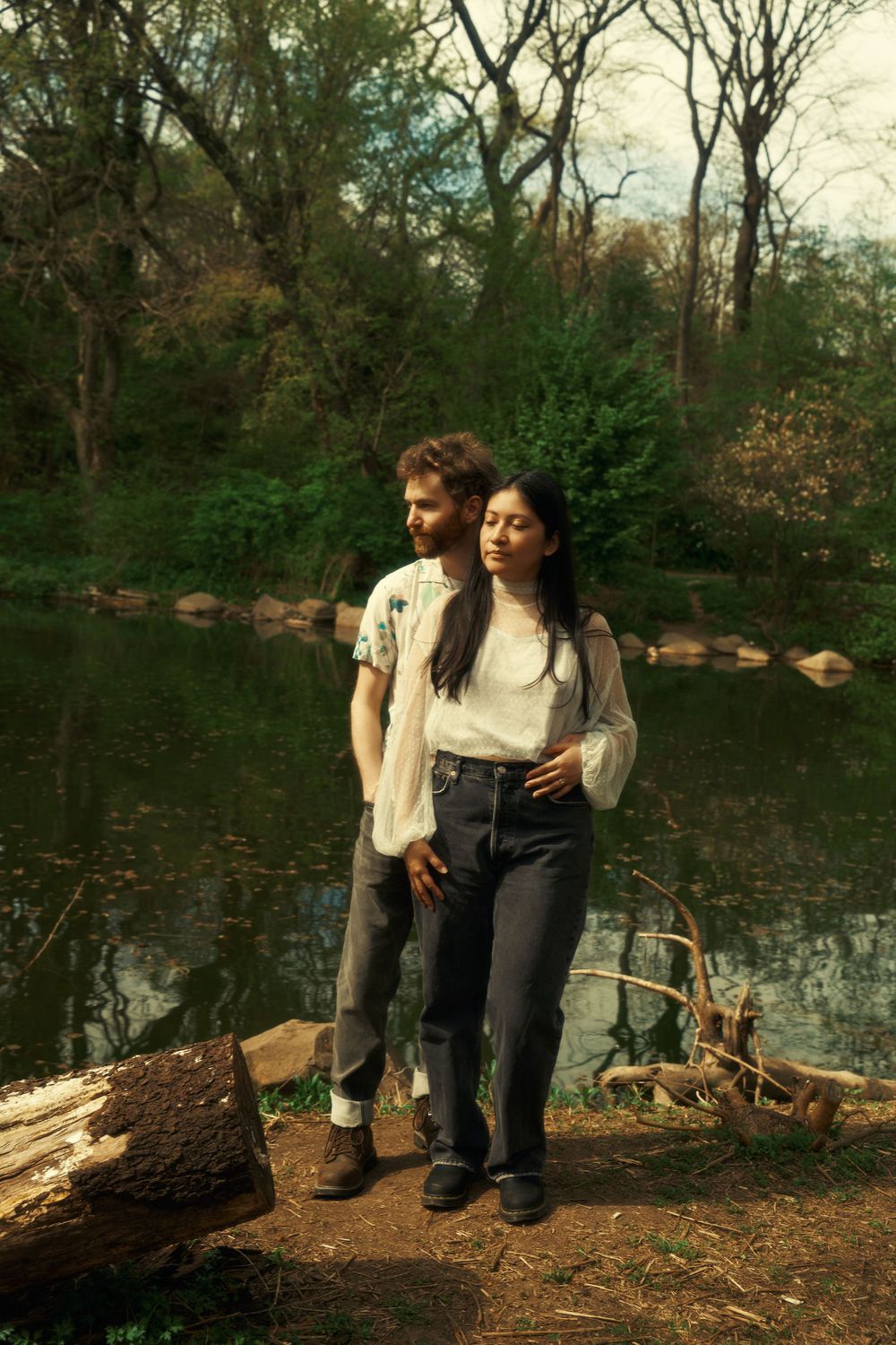A couple stands together near a serene pond with rocks and bare trees in the background during autumn prospect park boat house