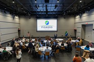 People gather at round tables in a conference room with a large screen displaying the Fishers logo on the wall.