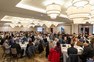 A large conference banquet hall filled with attendees seated at round tables under modern drum shade light fixtures.