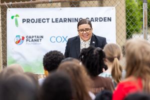 Speaker addresses crowd at Project Learning Garden event with outdoor banner display and palm tree logo visible in background.