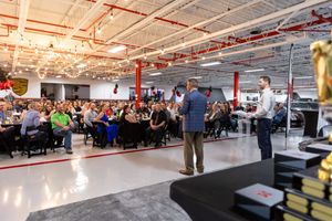 A speaker addresses a large crowd gathered in an industrial-style event space with exposed ceiling and red support beams.