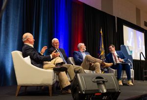 Panel discussion at business conference with four speakers seated in white chairs on stage with blue and red lighting.