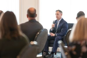 A speaker in a business suit sits on stage addressing an audience during a professional conference presentation.