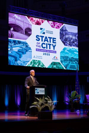 A speaker delivers remarks at a State of the City event with large projection screens displaying event branding.