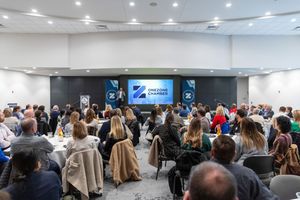 Large audience gathered in a modern conference room with white walls and blue digital screens during a corporate event.
