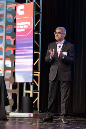A speaker gestures while presenting on stage at a corporate event with branded signage in the background.