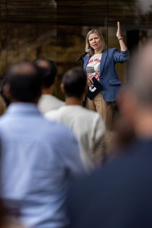 A speaker addresses a crowd while wearing a navy blazer and graphic t-shirt, gesturing with one hand raised.