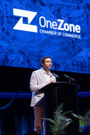 A speaker stands at a podium during an event with the OneZone Chamber of Commerce logo displayed on a blue backdrop.