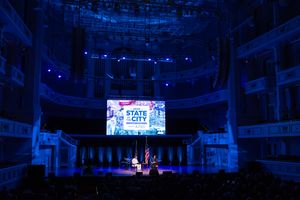 Concert stage illuminated with blue lighting and digital screens showing event details in a large indoor venue.