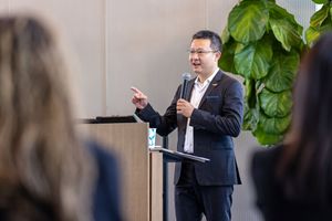 Professional speaker presenting from a podium in front of a large indoor plant during a business event.